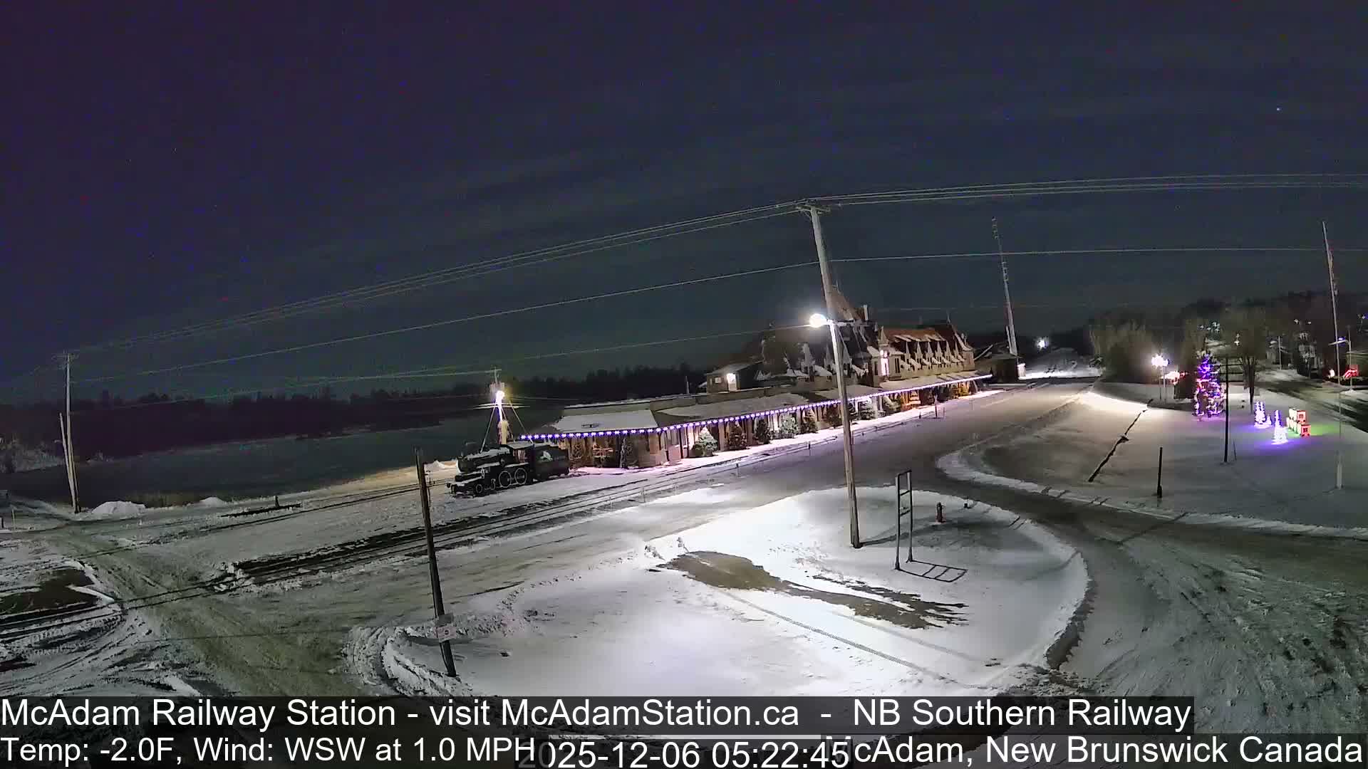 A snow-covered railway station, adorned with numerous festive lights, stands illuminated at night beside a track with a parked train engine, under a clear, cold sky, with additional decorated Christmas trees and figures nearby.