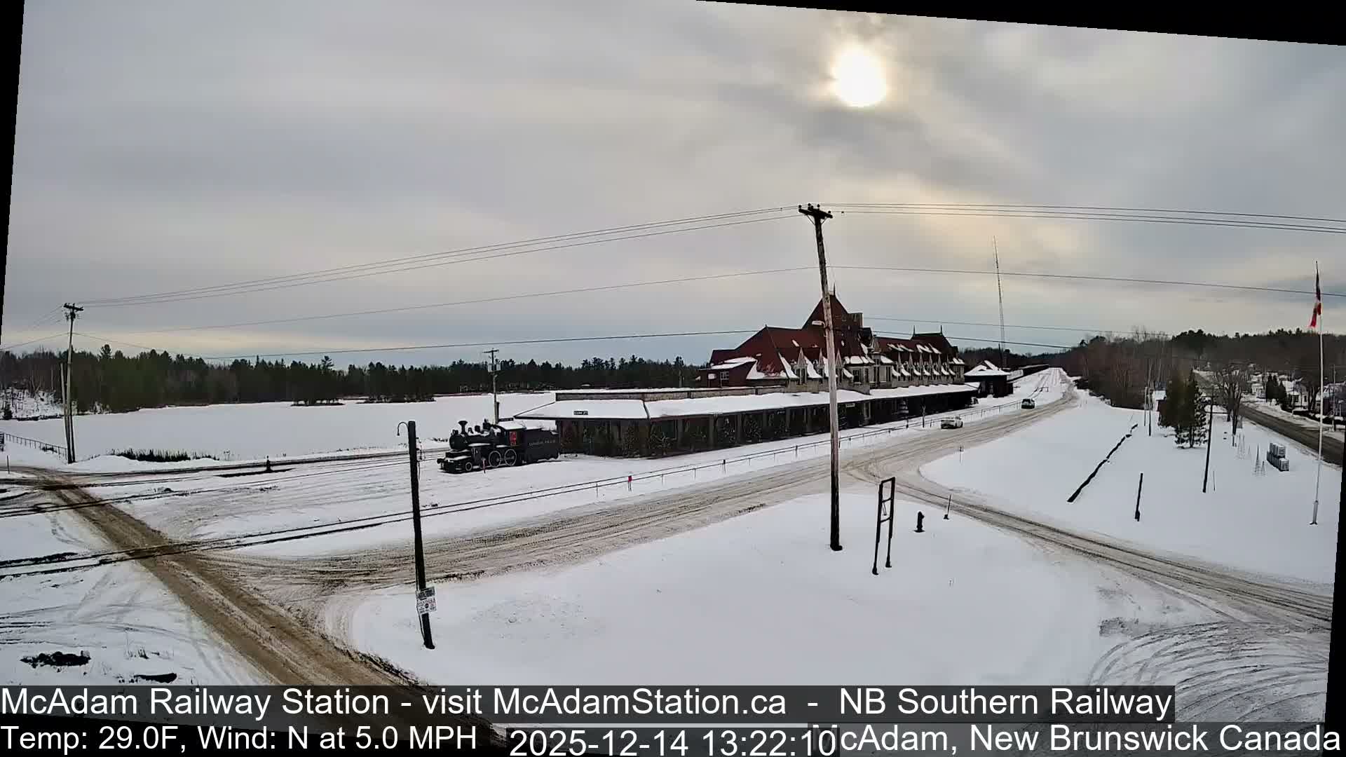 A snow-covered railway station, adorned with numerous festive lights, stands illuminated at night beside a track with a parked train engine, under a clear, cold sky, with additional decorated Christmas trees and figures nearby.