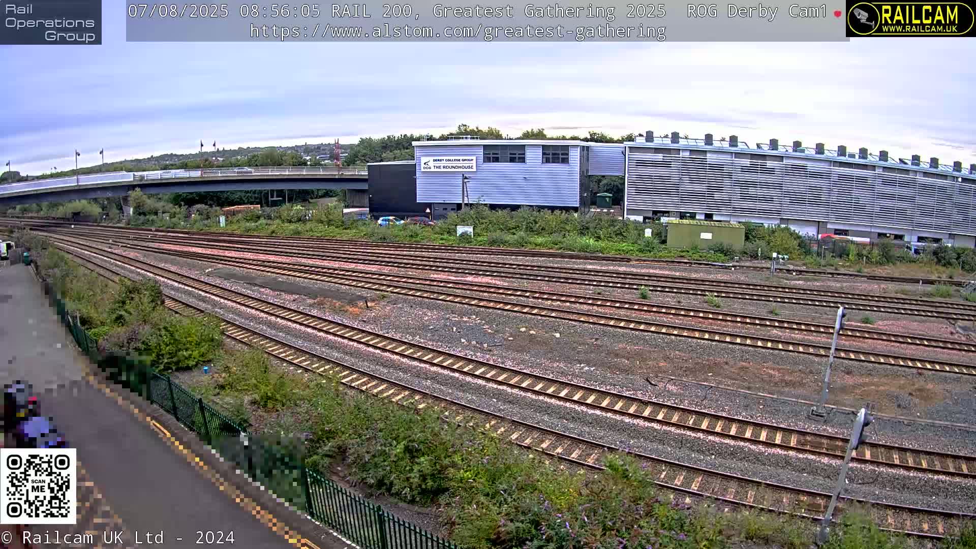 A high-angle, outdoor view on an overcast day shows many parallel railroad tracks next to a modern building and a bridge in the distance.