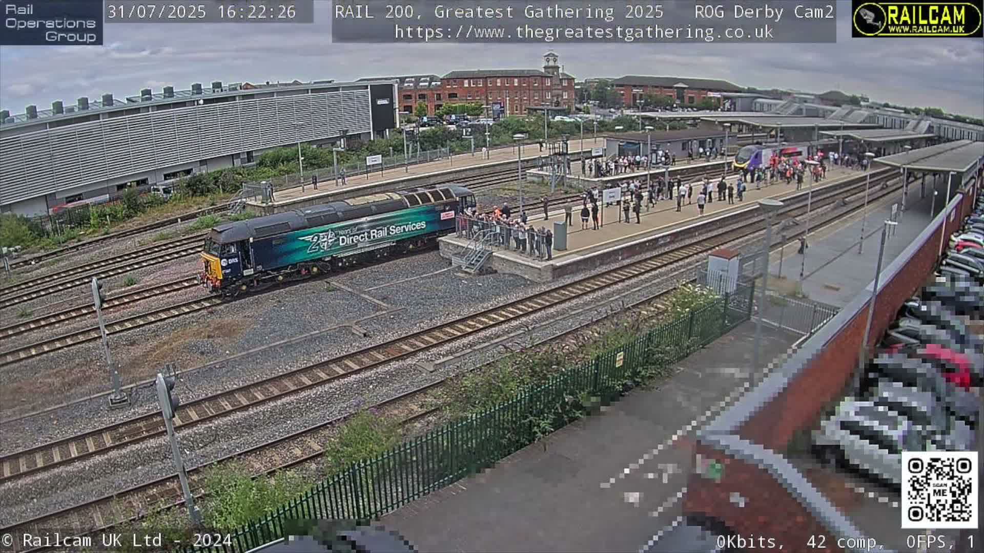 Under an overcast sky, a blue and green train is seen on tracks beside a bustling station platform crowded with people, while industrial buildings and a packed car park complete the scene.