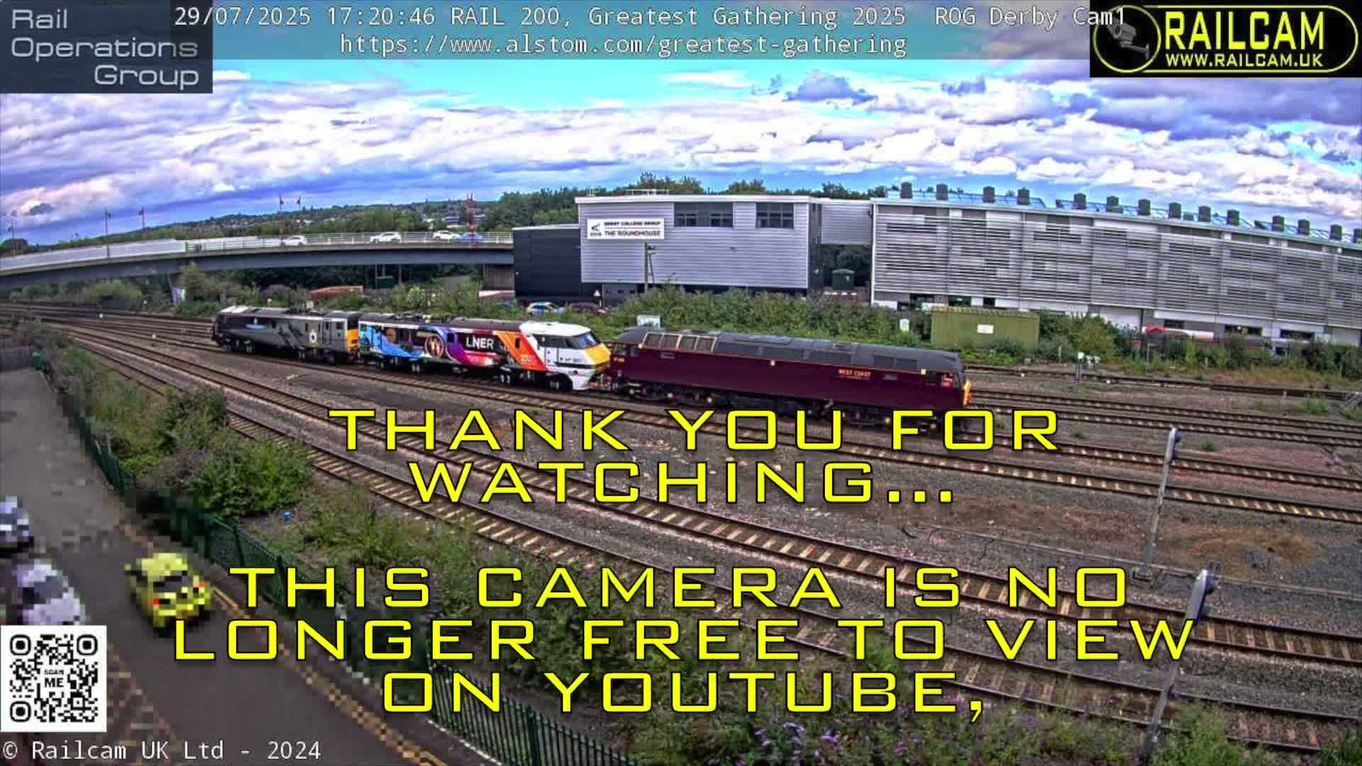 Multiple trains, including a dark red locomotive and a colorful LNER-branded carriage, rest on parallel tracks with lush greenery alongside, overlooked by a large grey building and a road bridge under a partly cloudy sky.