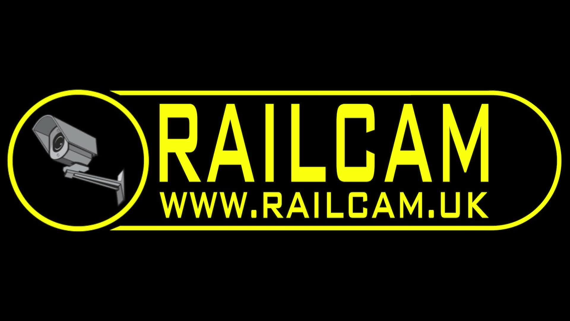 Multiple trains, including a dark red locomotive and a colorful LNER-branded carriage, rest on parallel tracks with lush greenery alongside, overlooked by a large grey building and a road bridge under a partly cloudy sky.