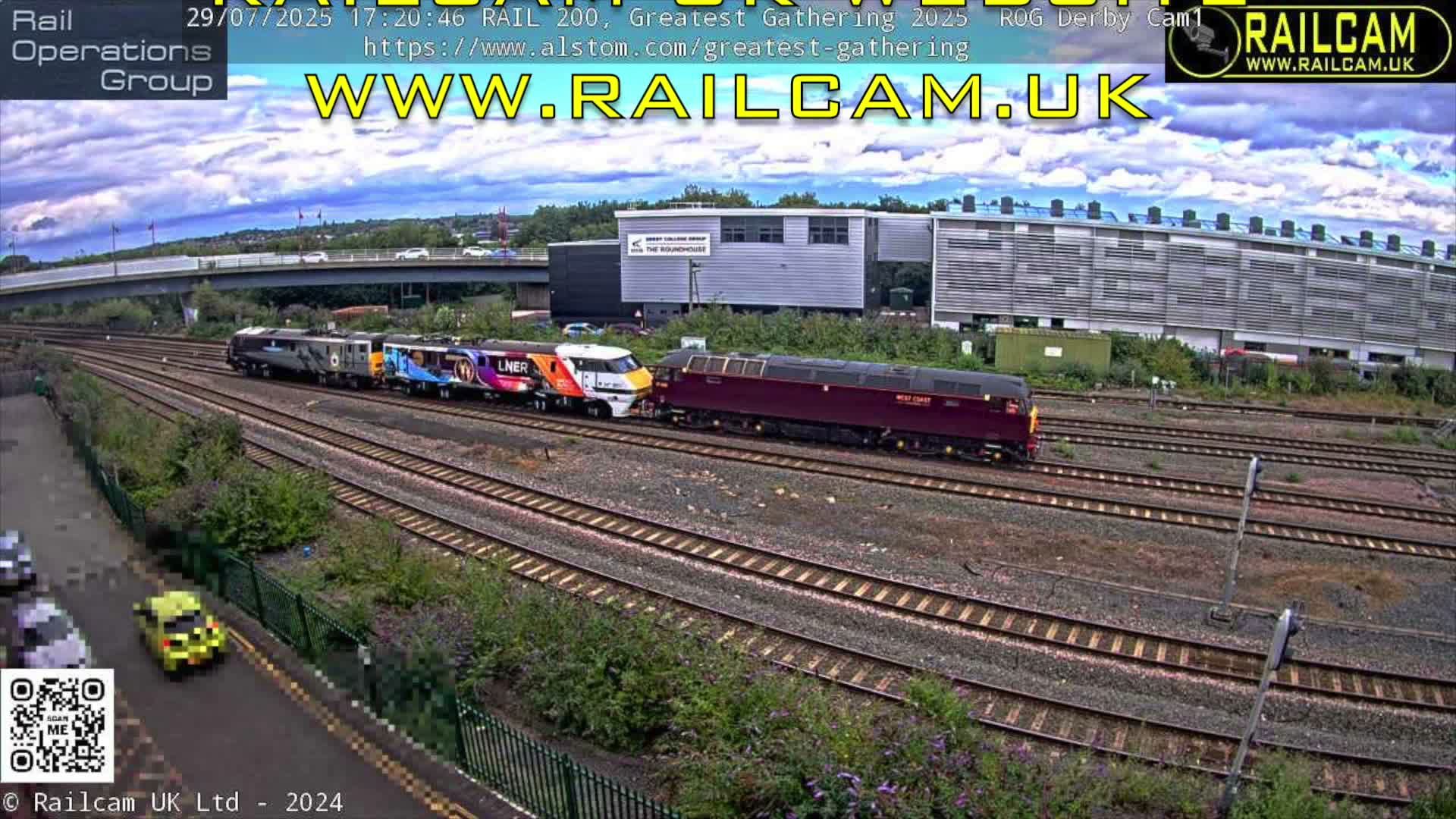 A colorful LNER train and a maroon locomotive are seen on parallel railway tracks, flanked by industrial buildings and a road bridge, under a partly cloudy sky.