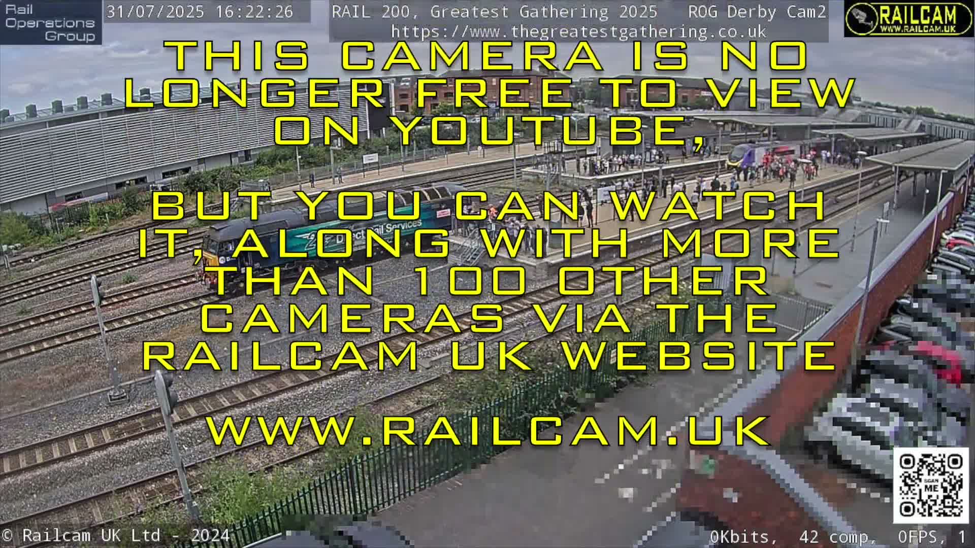 A colorful LNER train and a maroon locomotive are seen on parallel railway tracks, flanked by industrial buildings and a road bridge, under a partly cloudy sky.