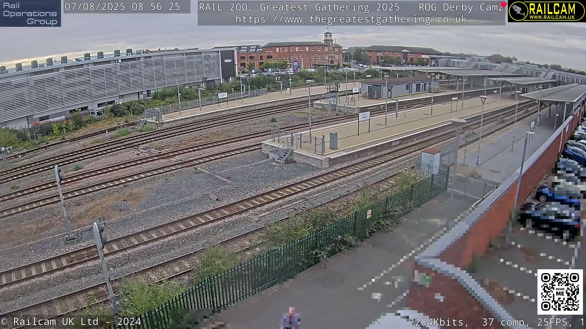 A high-angle, daytime view of a train station with multiple tracks, platforms, and surrounding buildings under partly cloudy skies.