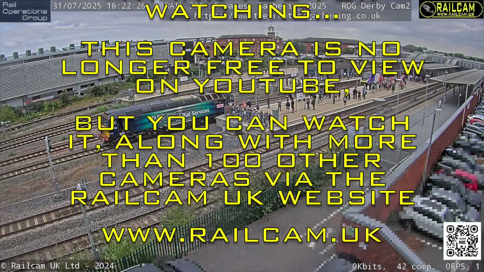 An elevated view captures an outdoor train station with a green and blue train stopped at a platform where numerous people are gathered, alongside multiple tracks, a large building in the background, and a packed parking lot, all under an overcast sky.