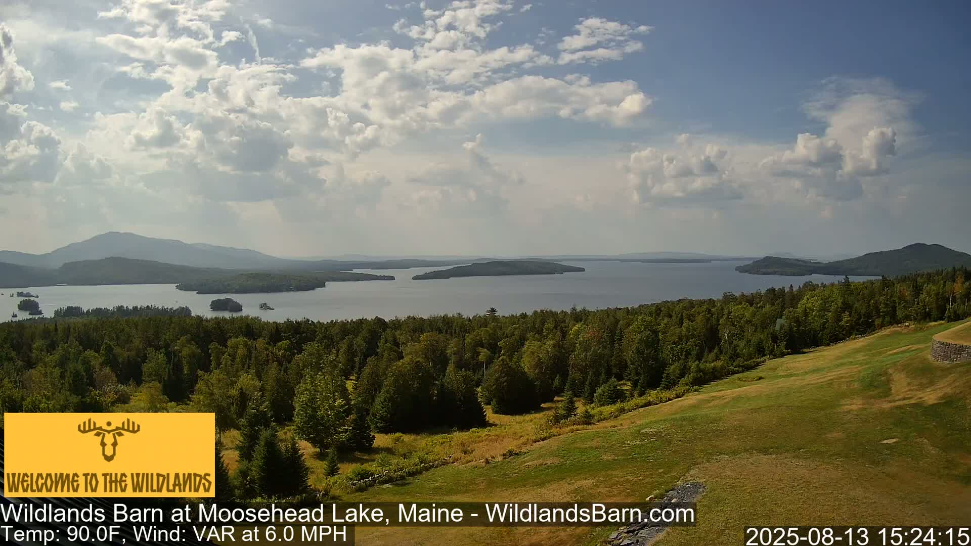 A panoramic view shows a large lake with islands, surrounded by green forested hills under a partly cloudy sky.