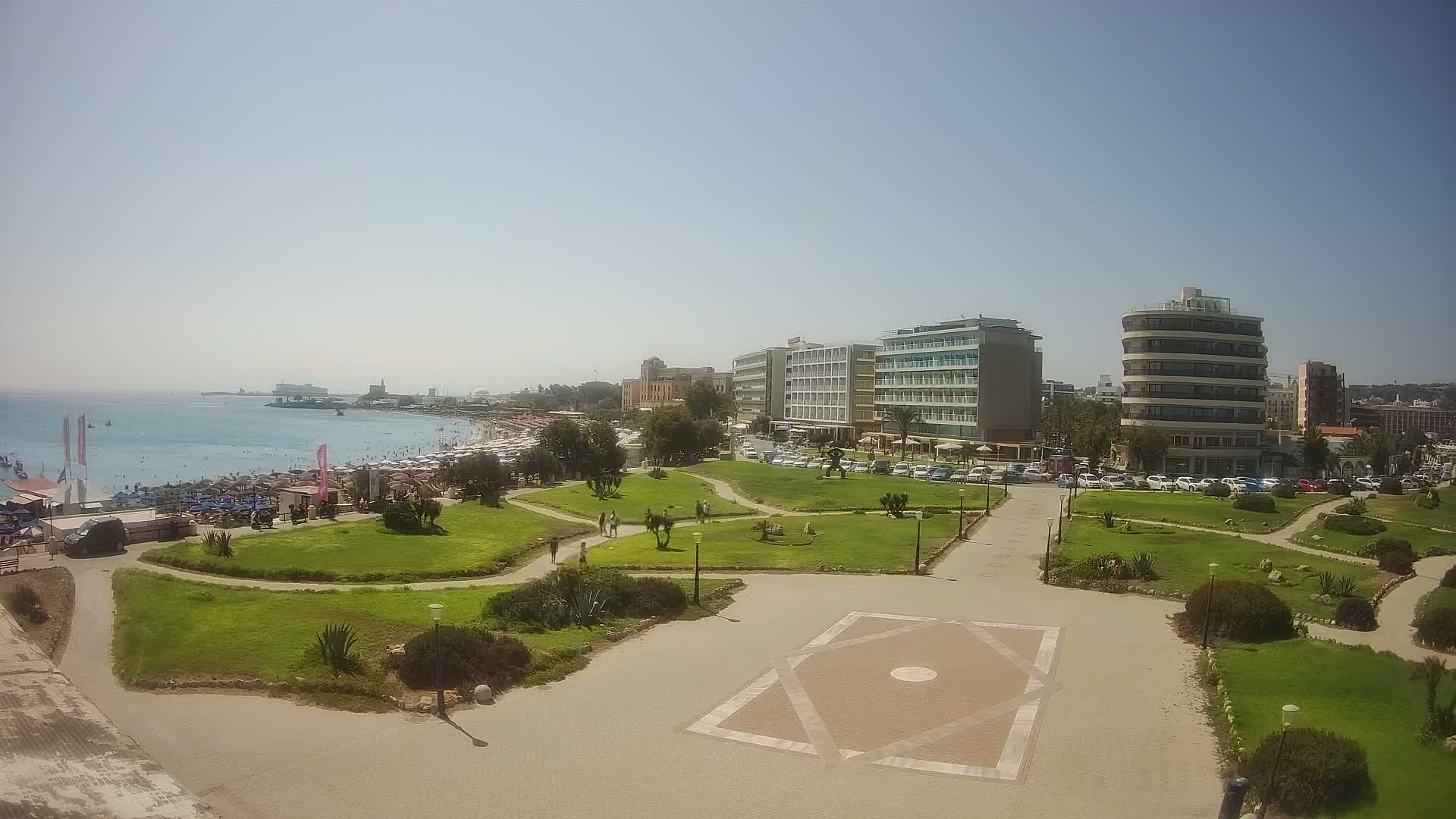 A sunny day reveals a coastal scene with a park featuring manicured lawns and pathways, overlooking a beach crowded with umbrellas and people, and several hotels and buildings along the waterfront.