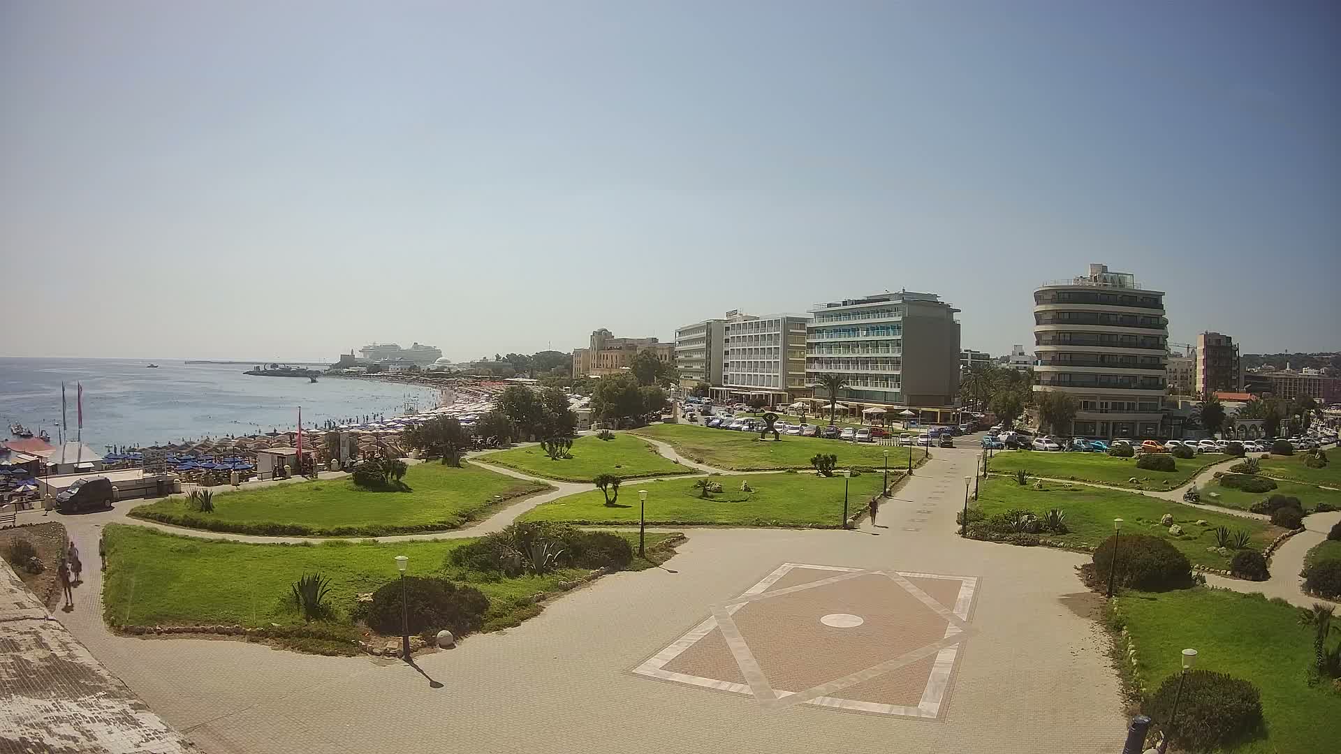A sunny day shows a seaside park with manicured lawns, paved walkways, and several hotels and buildings behind a crowded beach.