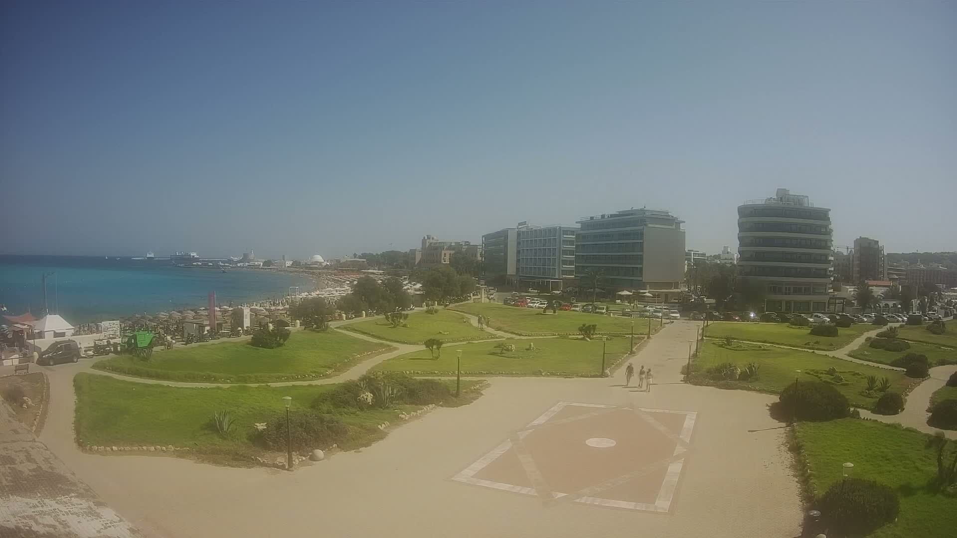 A sunny day shows a seaside park with manicured lawns, paved walkways, and several hotels and buildings behind a crowded beach.
