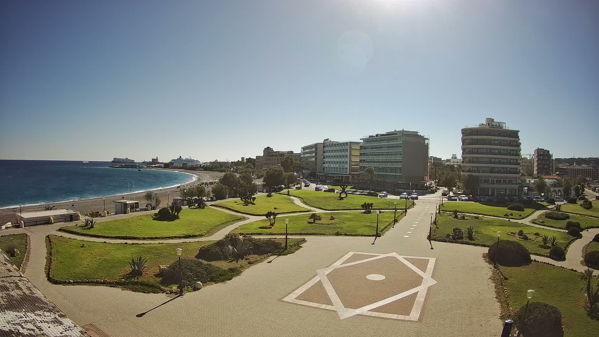 A sunny day illuminates a wide coastal city view featuring a long pebbled beach next to the deep blue sea with distant ships, a large green park with paved paths and an octagonal star design in the foreground, and modern buildings under a clear blue sky.