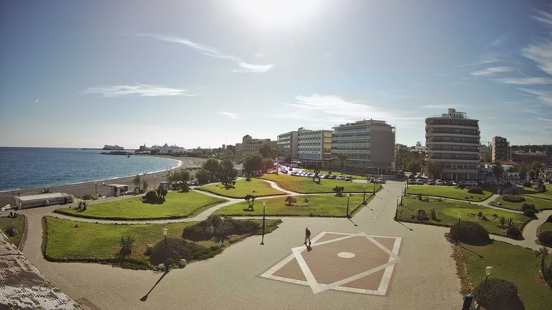 Under a bright, sunny sky with scattered wispy clouds, this elevated view showcases a calm blue sea and pebble beach on the left, an expansive green urban park with a person walking in the center, and a city skyline featuring a mix of modern and older buildings to the right.