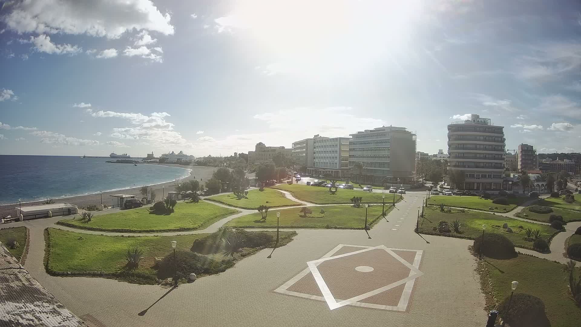On a bright, partly cloudy day, a coastal city scene unfolds with a wide beach and calm blue sea dotted with ships, leading to green parks, paved walkways with a geometric pattern, and numerous modern multi-story buildings alongside busy roads.