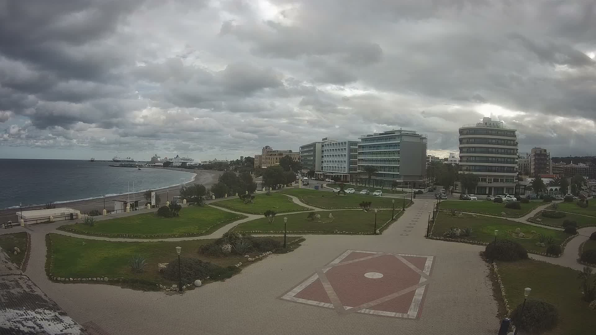 On a bright, partly cloudy day, a coastal city scene unfolds with a wide beach and calm blue sea dotted with ships, leading to green parks, paved walkways with a geometric pattern, and numerous modern multi-story buildings alongside busy roads.