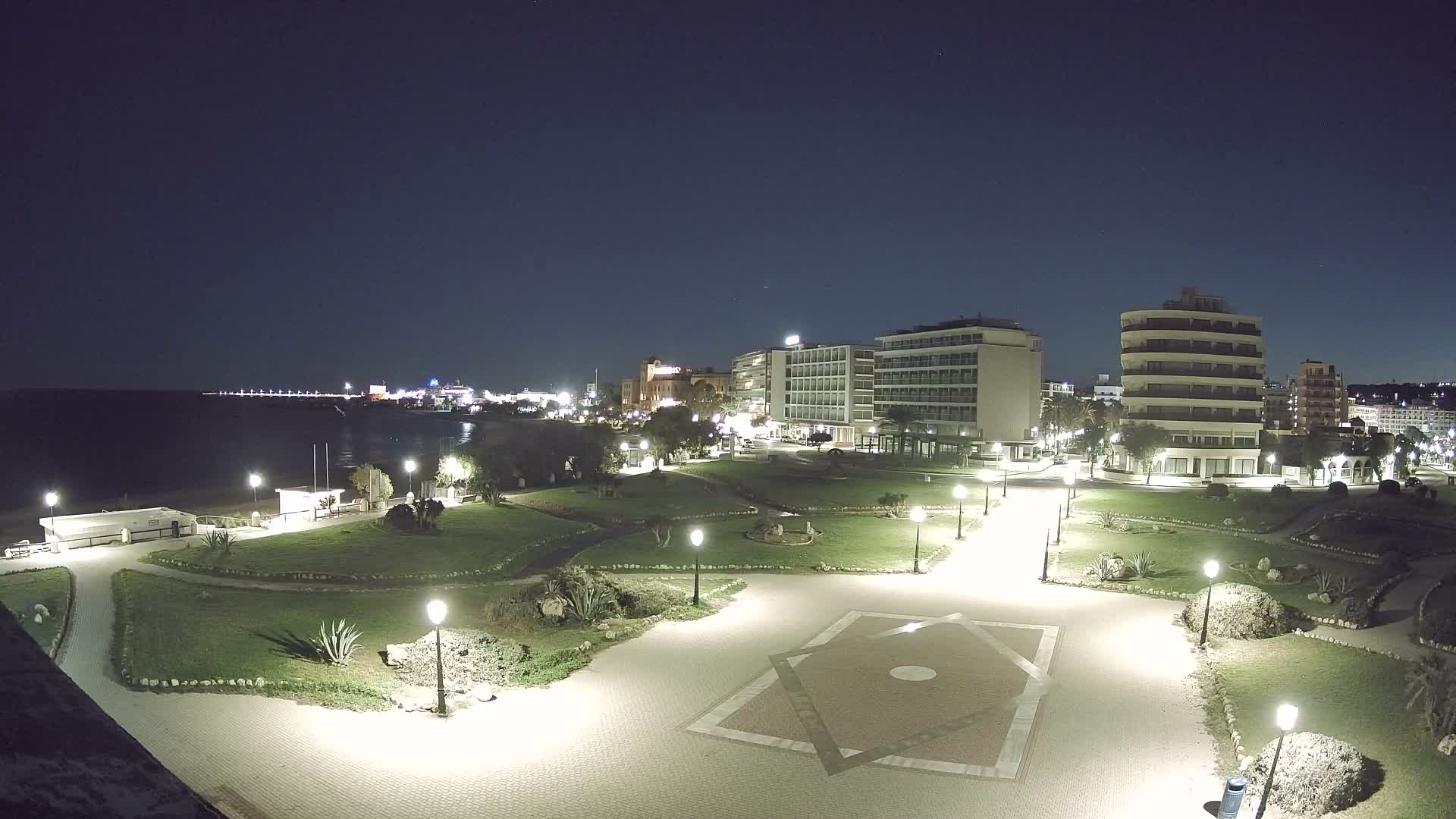 A clear, starry night reveals a brightly lit coastal city featuring a tranquil sea, a green park with paved paths, and numerous illuminated buildings along the shoreline.