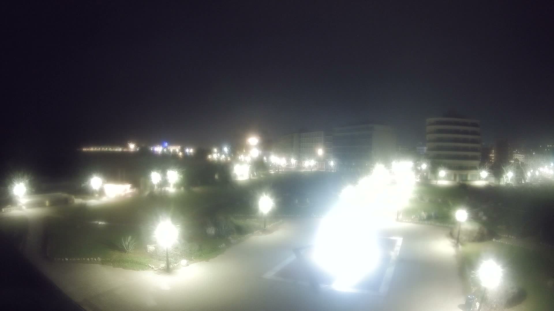 A clear, starry night reveals a brightly lit coastal city featuring a tranquil sea, a green park with paved paths, and numerous illuminated buildings along the shoreline.