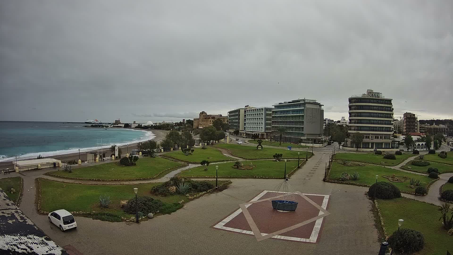 Under a gray, overcast sky, a coastal city scene features a green park with winding paths and a geometrically paved plaza in the foreground, leading to a turquoise beach with gentle waves and a distant ferry, all backed by a mix of modern and traditional multi-story buildings.