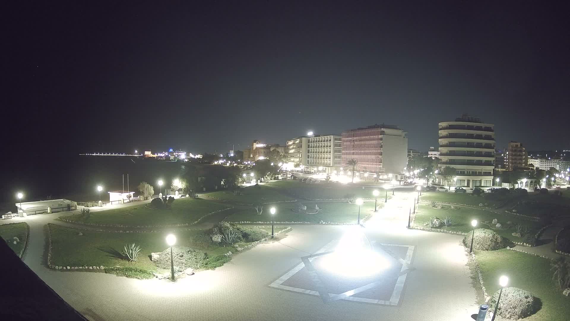 Under a gray, overcast sky, a coastal city scene features a green park with winding paths and a geometrically paved plaza in the foreground, leading to a turquoise beach with gentle waves and a distant ferry, all backed by a mix of modern and traditional multi-story buildings.