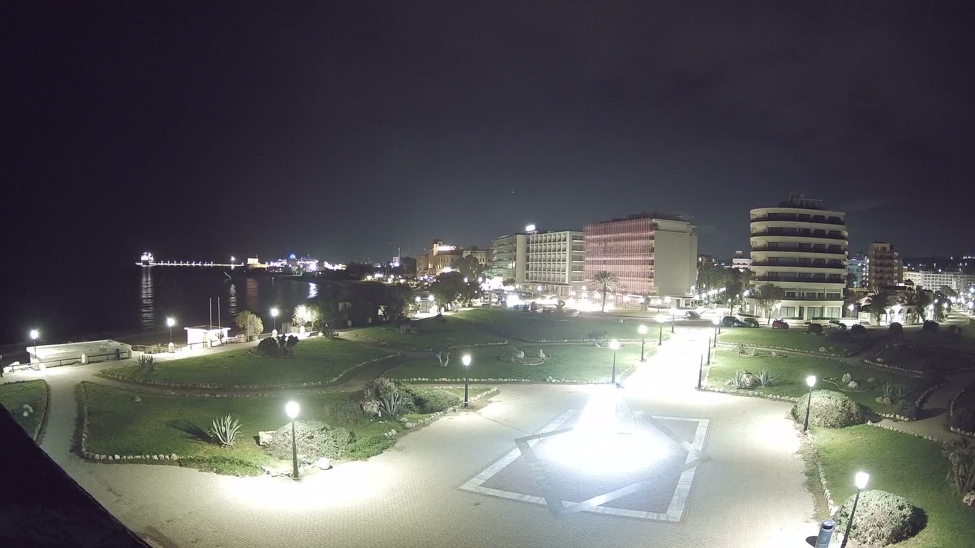 Under a gray, overcast sky, a coastal city scene features a green park with winding paths and a geometrically paved plaza in the foreground, leading to a turquoise beach with gentle waves and a distant ferry, all backed by a mix of modern and traditional multi-story buildings.