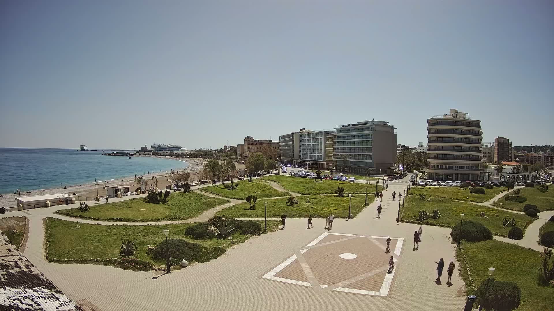 Under a gray, overcast sky, a coastal city scene features a green park with winding paths and a geometrically paved plaza in the foreground, leading to a turquoise beach with gentle waves and a distant ferry, all backed by a mix of modern and traditional multi-story buildings.