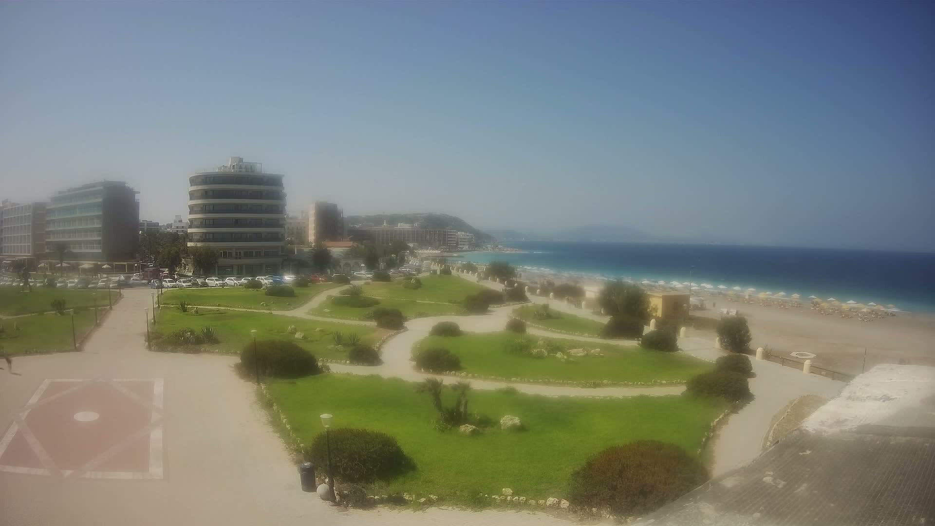 A well-maintained park with winding paths overlooks a sandy beach and calm ocean under a clear, sunny sky, with several buildings visible in the background.
