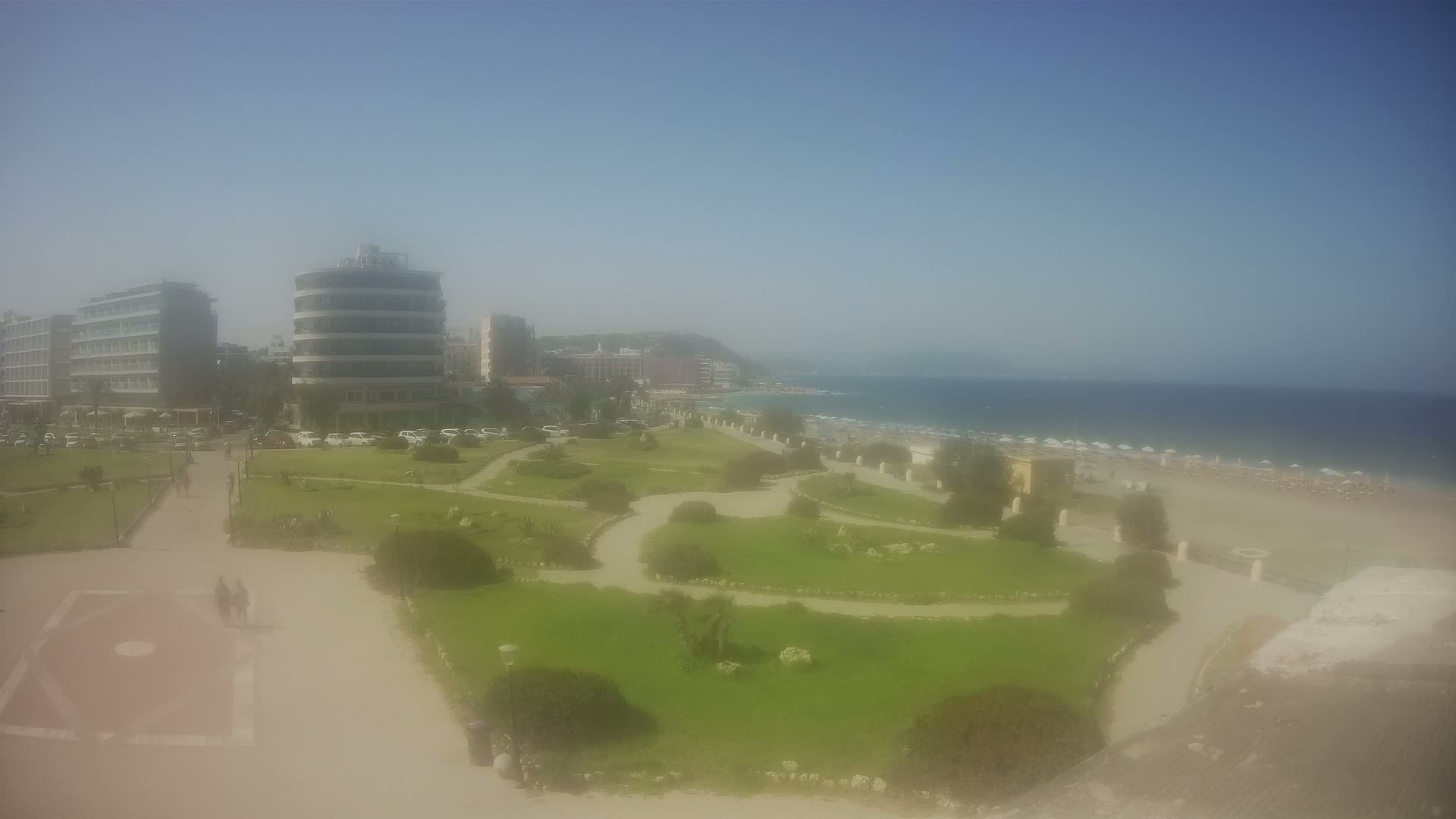 A seaside park with winding paths and manicured lawns overlooks a sandy beach with umbrellas under a clear, sunny sky.