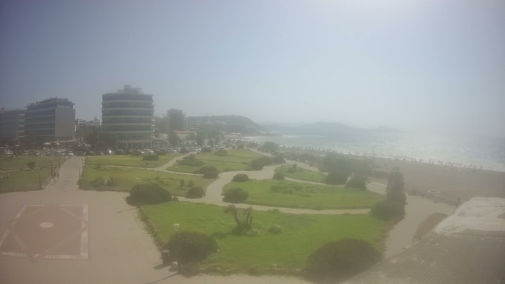 A seaside park with winding paths and manicured lawns overlooks a sandy beach with umbrellas under a clear, sunny sky.
