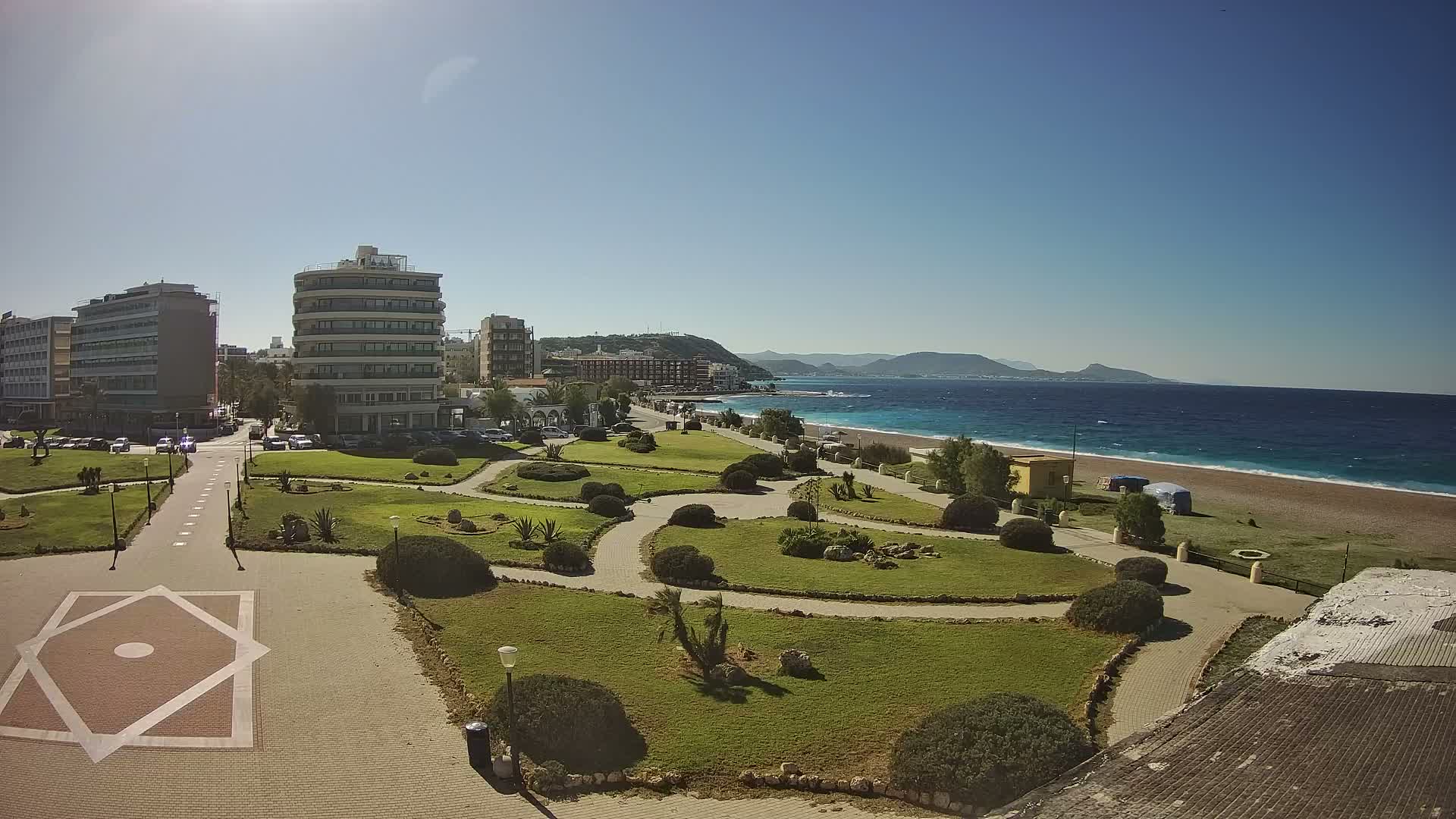 On a bright, clear day, an expansive coastal view presents a green park with winding paths, urban buildings, a pebble beach, the deep blue sea, and distant mountains under a brilliant blue sky.