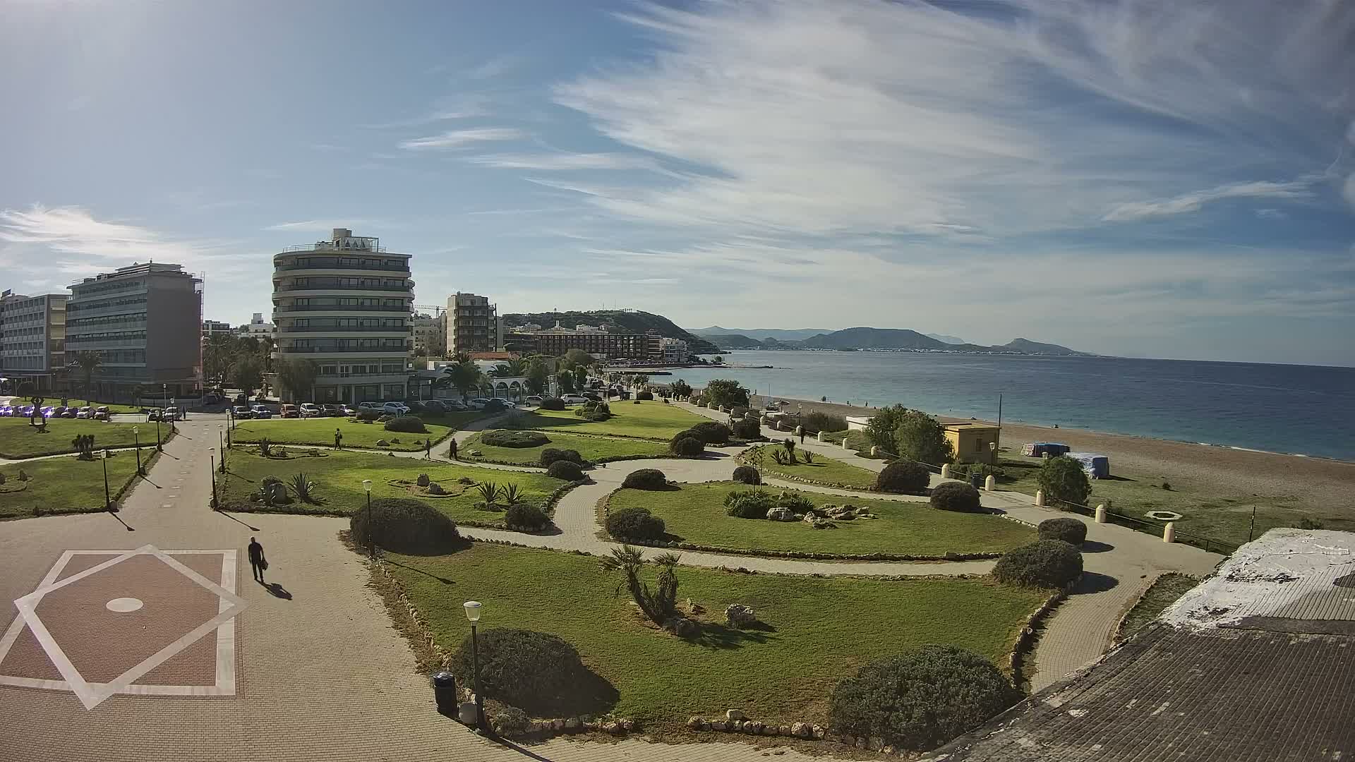 A bright and partly cloudy day reveals a coastal city with modern buildings overlooking a green park with winding paths, a sandy beach, and the calm blue sea stretching towards distant mountains.