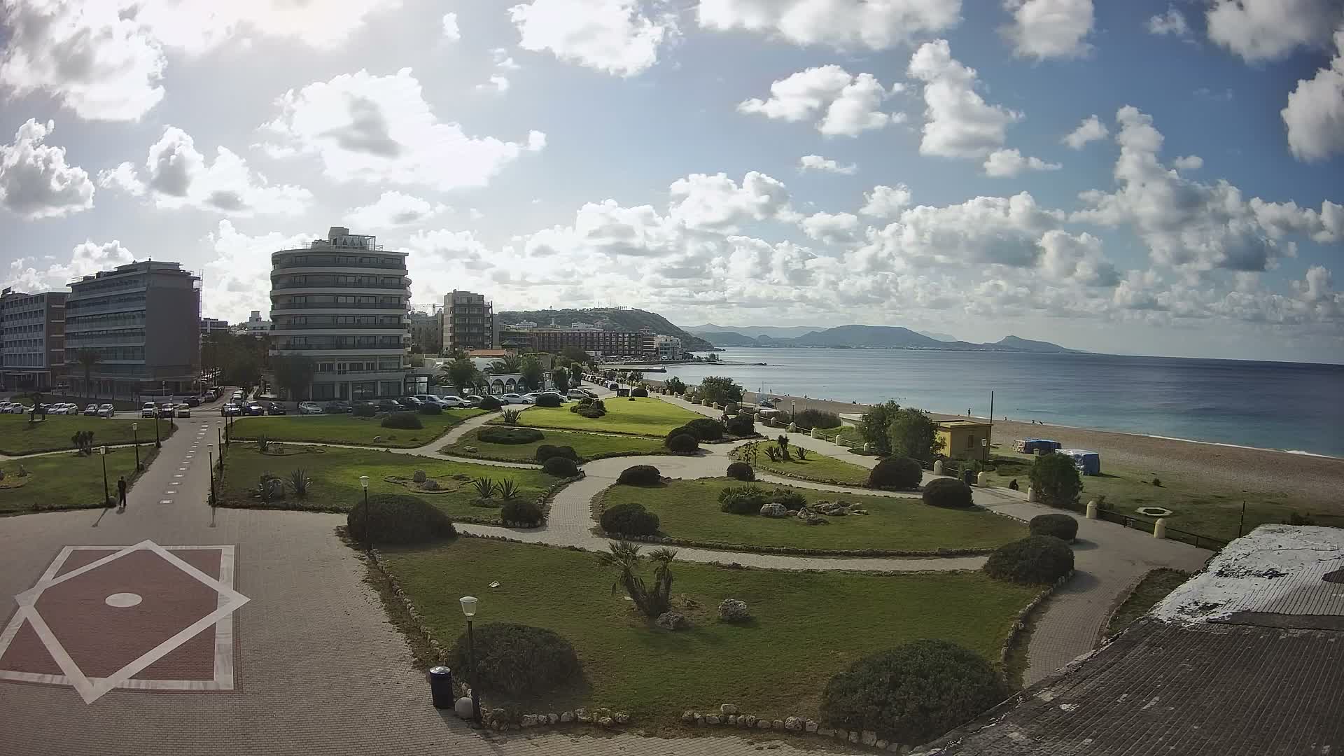 A bright and partly cloudy day reveals a coastal city with modern buildings overlooking a green park with winding paths, a sandy beach, and the calm blue sea stretching towards distant mountains.