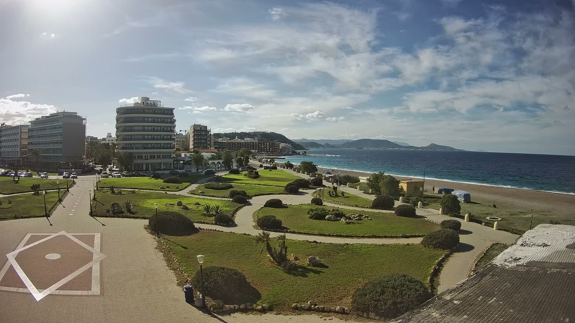 The image captures a panoramic view of a coastal city featuring modern buildings, a landscaped park with winding paths and green spaces, a pebbly beach, and the blue sea with distant mountains under a bright, partly cloudy sky.