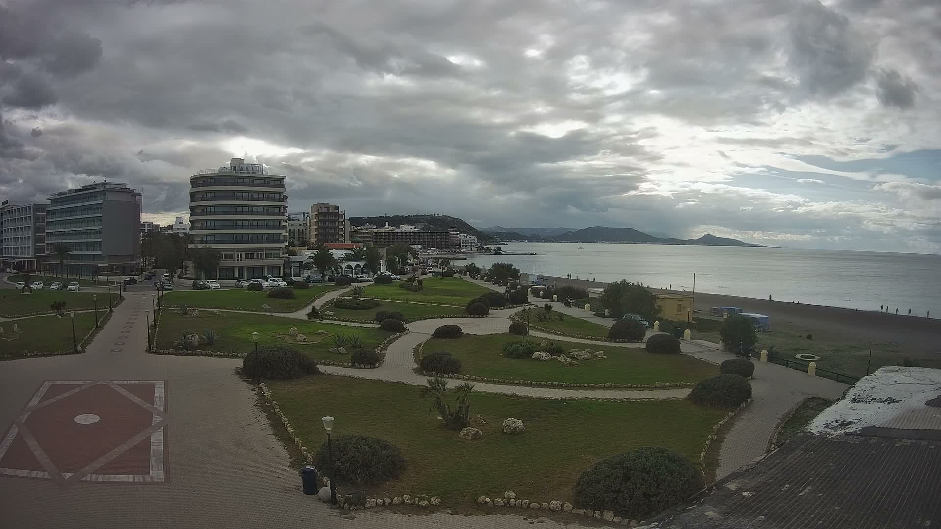 The image captures a panoramic view of a coastal city featuring modern buildings, a landscaped park with winding paths and green spaces, a pebbly beach, and the blue sea with distant mountains under a bright, partly cloudy sky.