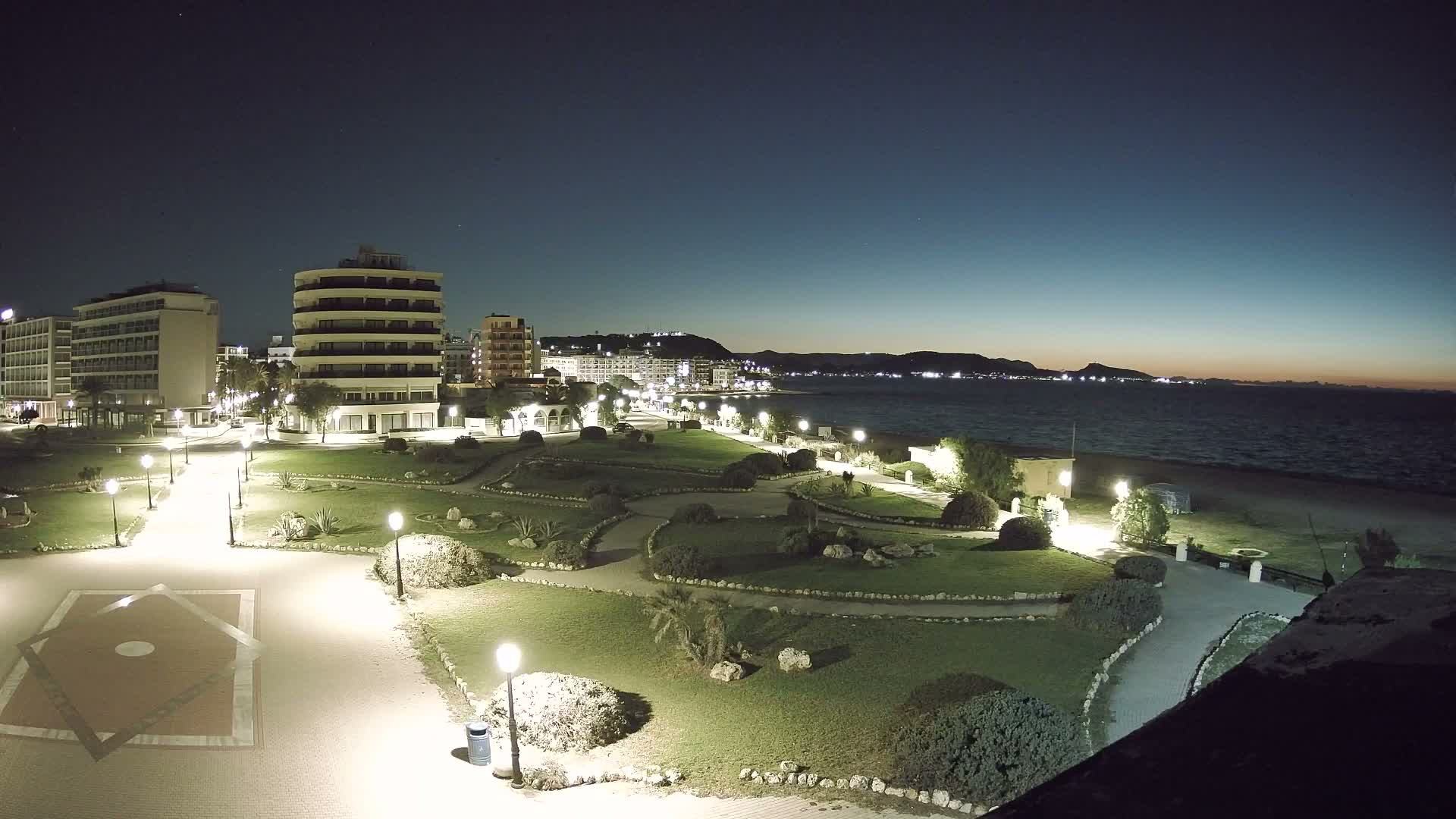 A clear night view shows an illuminated coastal park with winding paths and buildings, looking out towards the dark sea and a city-lit coastline backed by distant mountains.