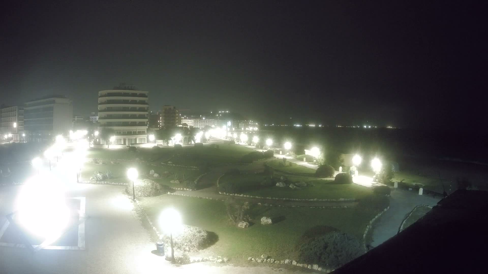 A clear night view shows an illuminated coastal park with winding paths and buildings, looking out towards the dark sea and a city-lit coastline backed by distant mountains.
