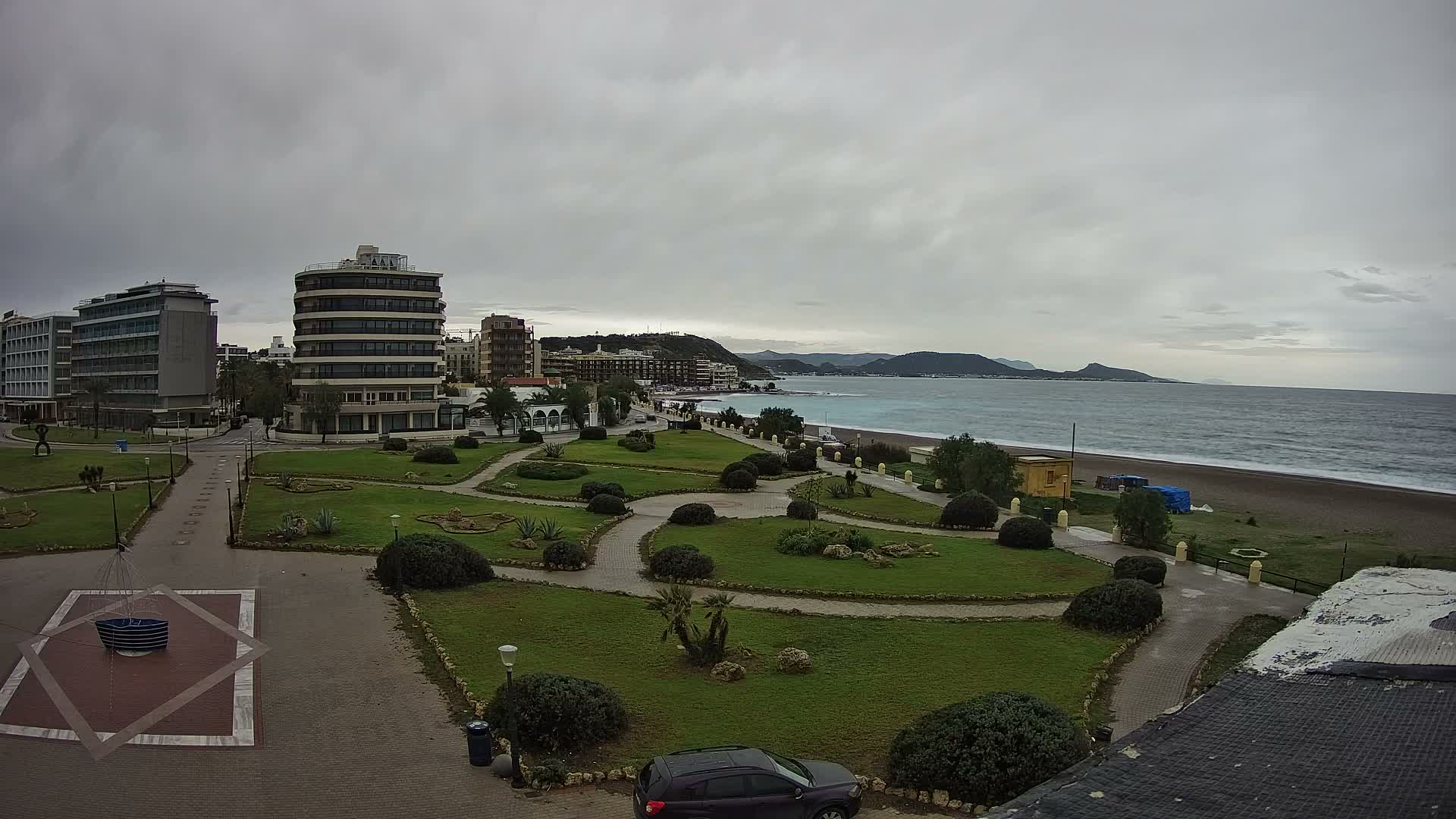 An overcast sky hangs over a coastal city scene, featuring a green park with winding paths and shrubs in the foreground, leading to a sandy beach, the sea, and distant hills lined with various buildings.
