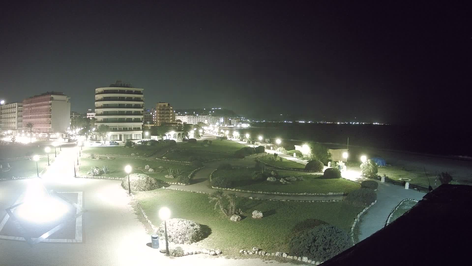 An overcast sky hangs over a coastal city scene, featuring a green park with winding paths and shrubs in the foreground, leading to a sandy beach, the sea, and distant hills lined with various buildings.