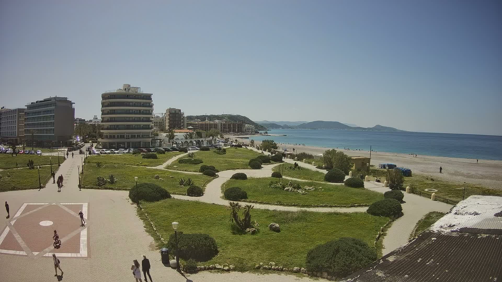 An overcast sky hangs over a coastal city scene, featuring a green park with winding paths and shrubs in the foreground, leading to a sandy beach, the sea, and distant hills lined with various buildings.