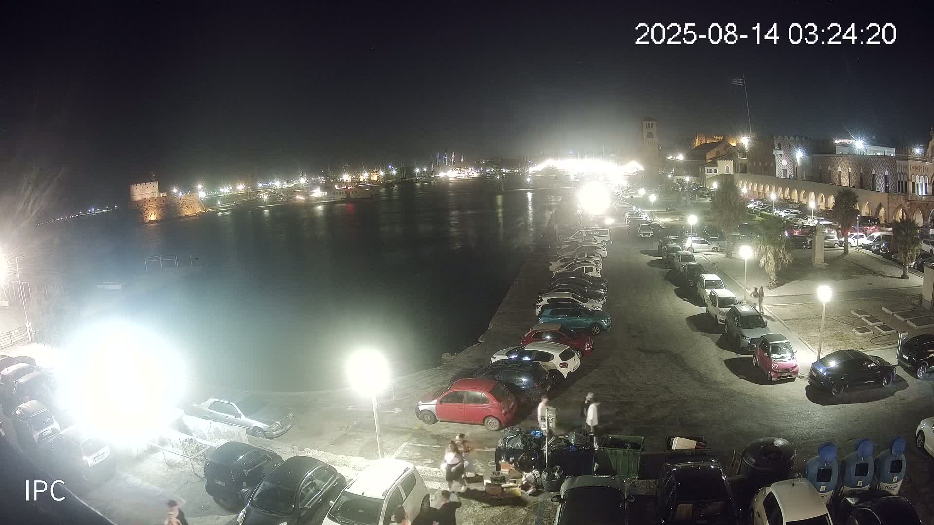 A nighttime view of a waterfront parking lot with cars parked along a seawall, under clear skies, overlooking a calm body of water with a stone fortress in the distance.