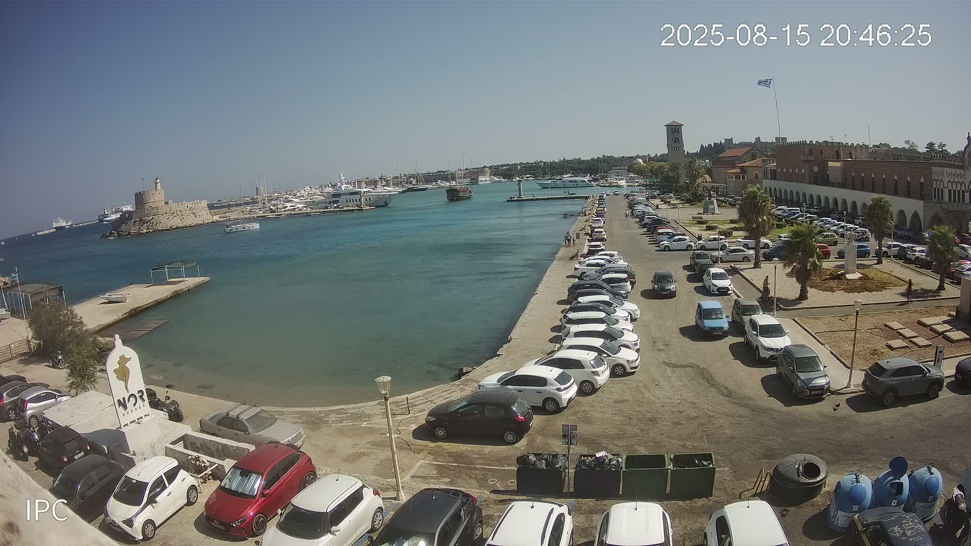 A harbor filled with boats and yachts under a sunny sky is seen from a high vantage point, with a parking lot and buildings lining the waterfront.