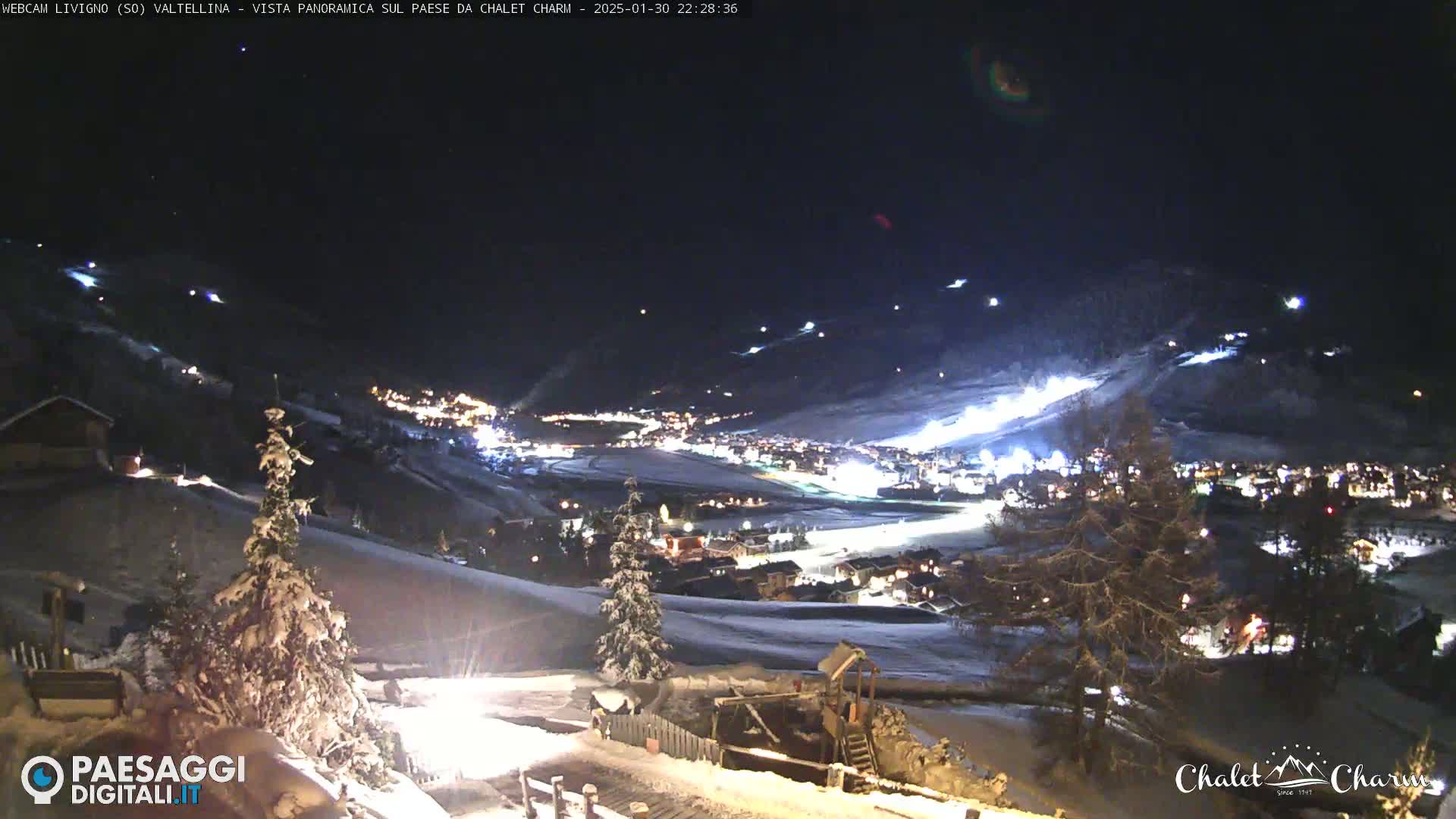 A snow-covered mountain village at night is illuminated, with lit ski slopes visible on the surrounding hills under a clear, dark sky.