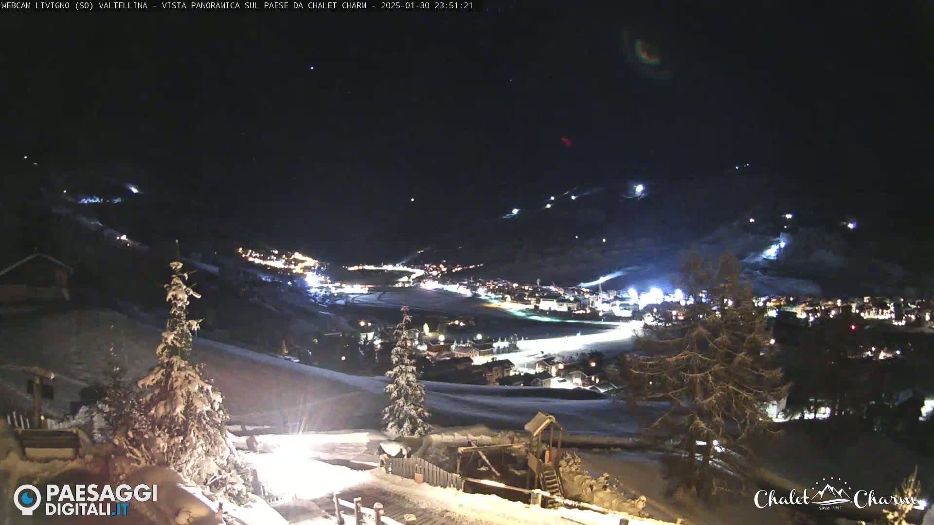 A snow-covered landscape at night shows a town illuminated below, with ski slopes visible on the surrounding hills under a clear, dark sky.