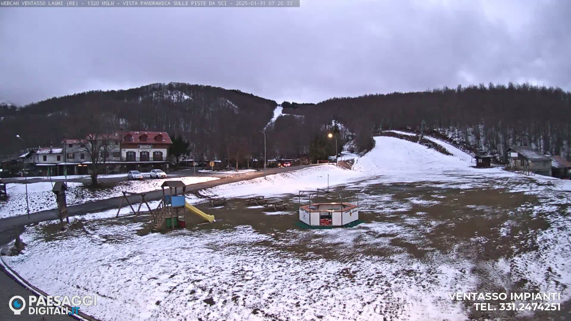 A snow-covered playground and parking lot sit below a ski slope on a hill, under a cloudy, overcast sky.