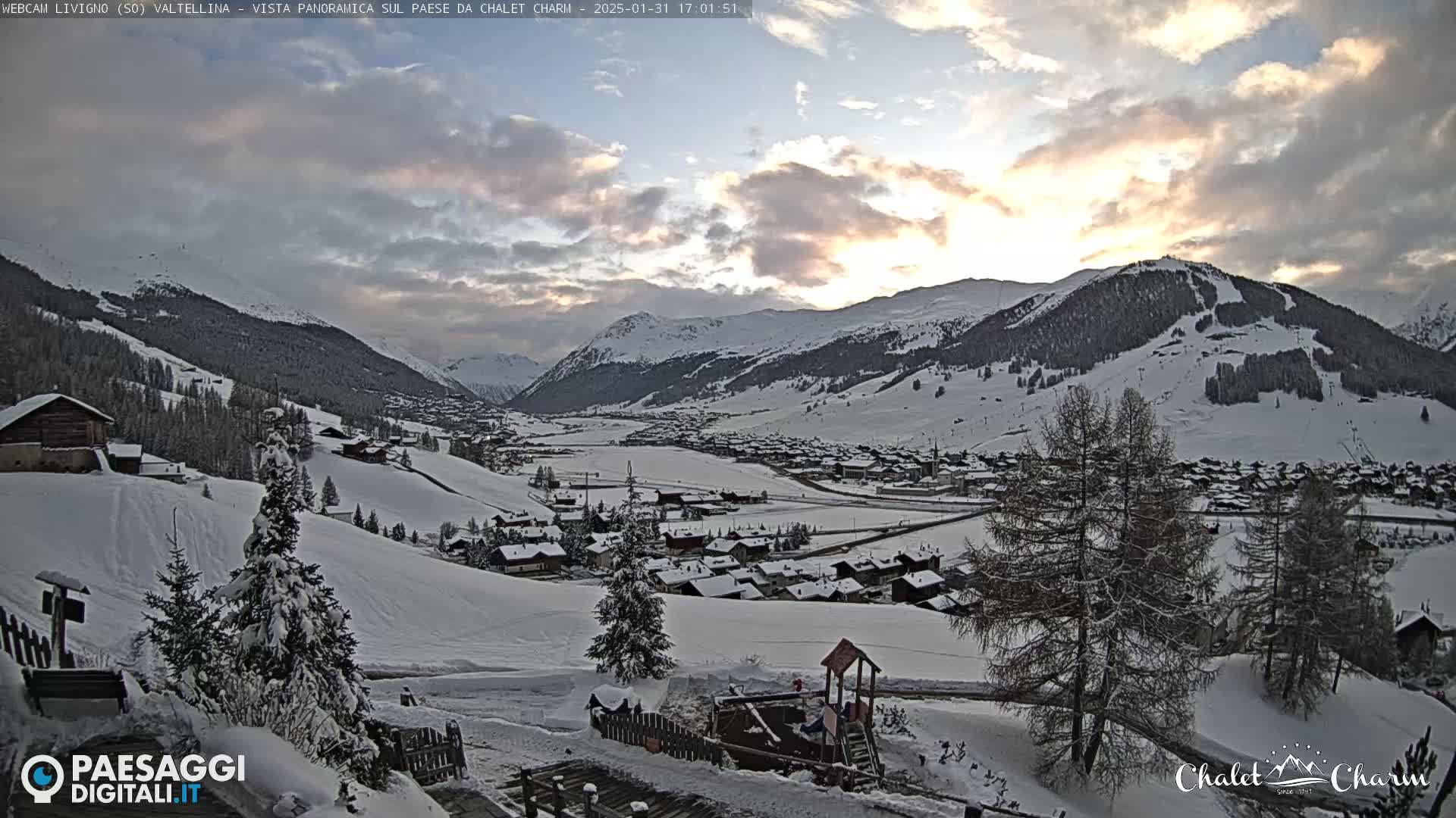 A snow-covered village nestled in a valley between snow-capped mountains under a partly cloudy sunset sky.