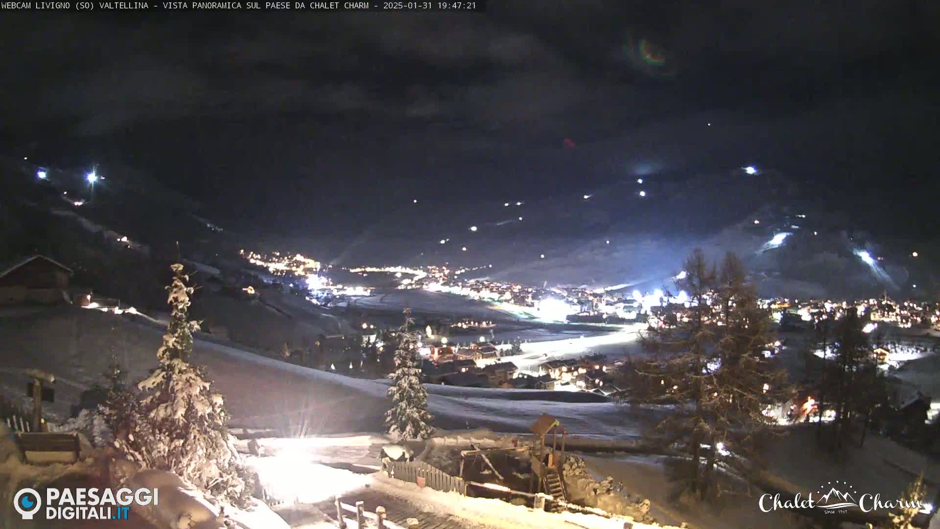A nighttime panoramic view of a snow-covered village illuminated by lights, with a cloudy sky and several streaks of light possibly from skiers or snowboarders on a nearby slope.