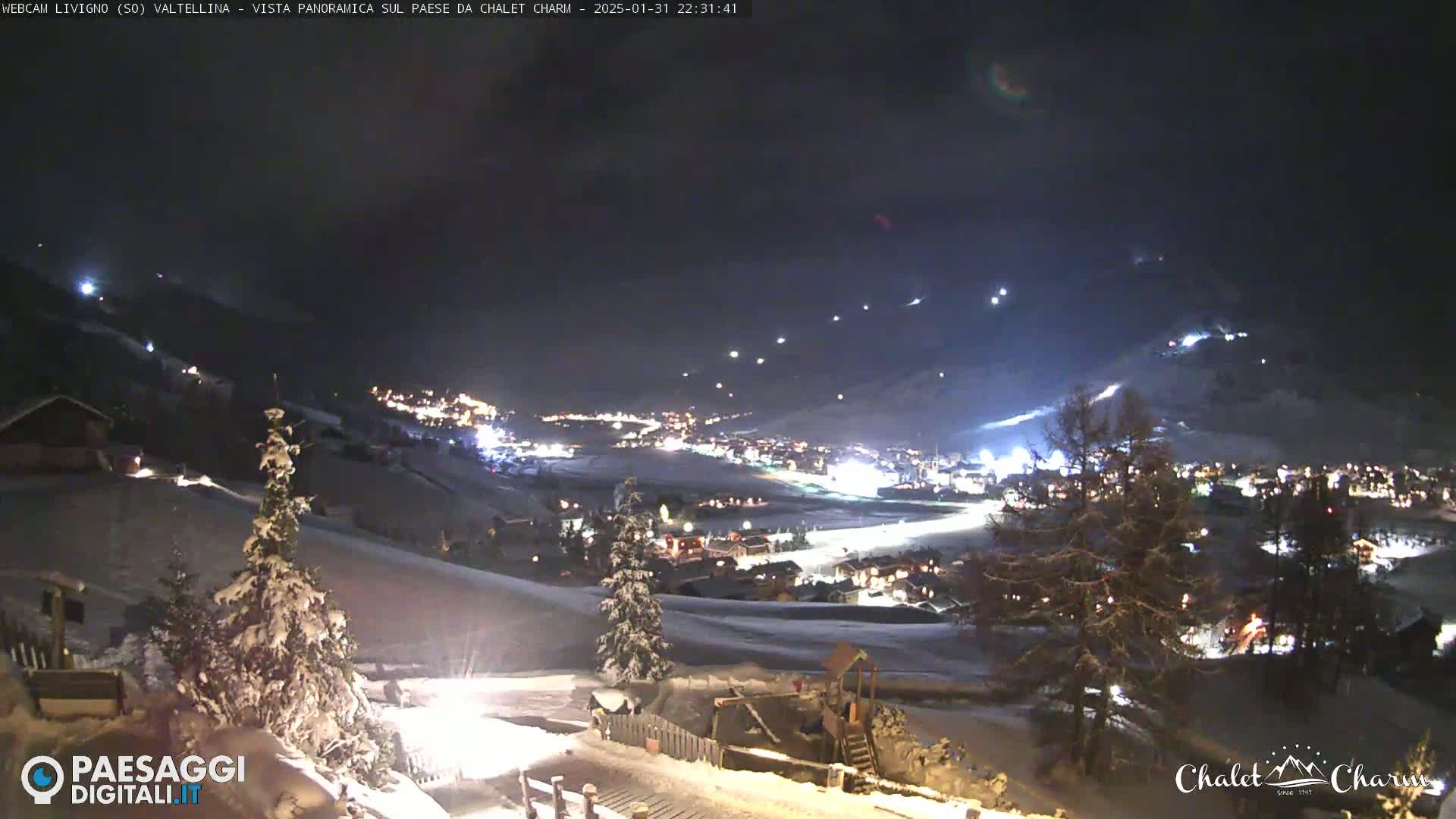 A snow-covered village at night is illuminated by lights, with a hillside showing signs of nighttime skiing activity under a dark sky.