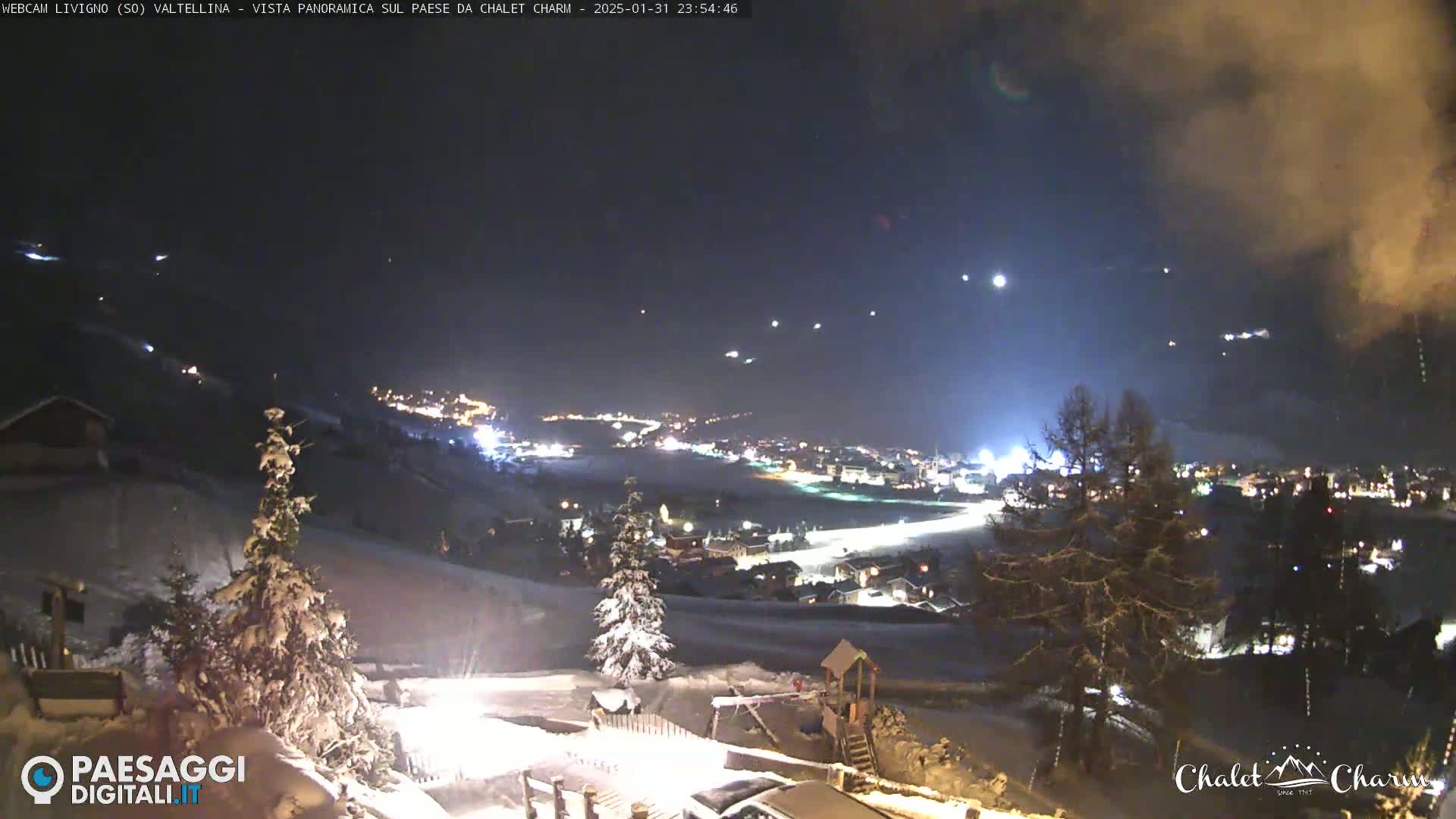 A snow-covered landscape at night, illuminated by the lights of a town nestled in a valley below, under a dark sky with some clouds and faint lights possibly from stars or aircraft.