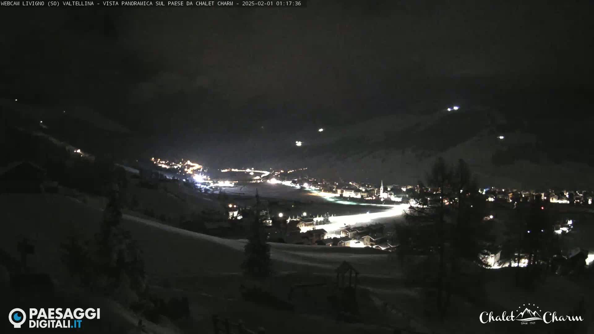 A nighttime view of a snow-covered town illuminated by lights, seen from a hilltop under a dark, clear sky.