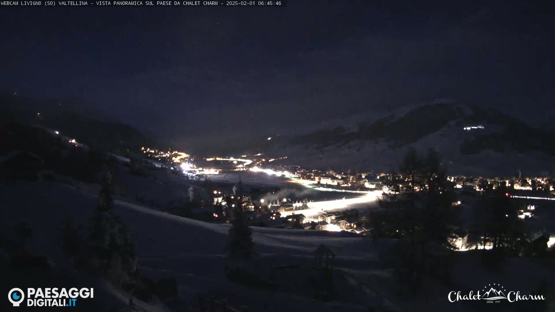 A snow-covered, nighttime landscape shows a village illuminated below, nestled in a valley between dark mountains under a clear, dark sky.