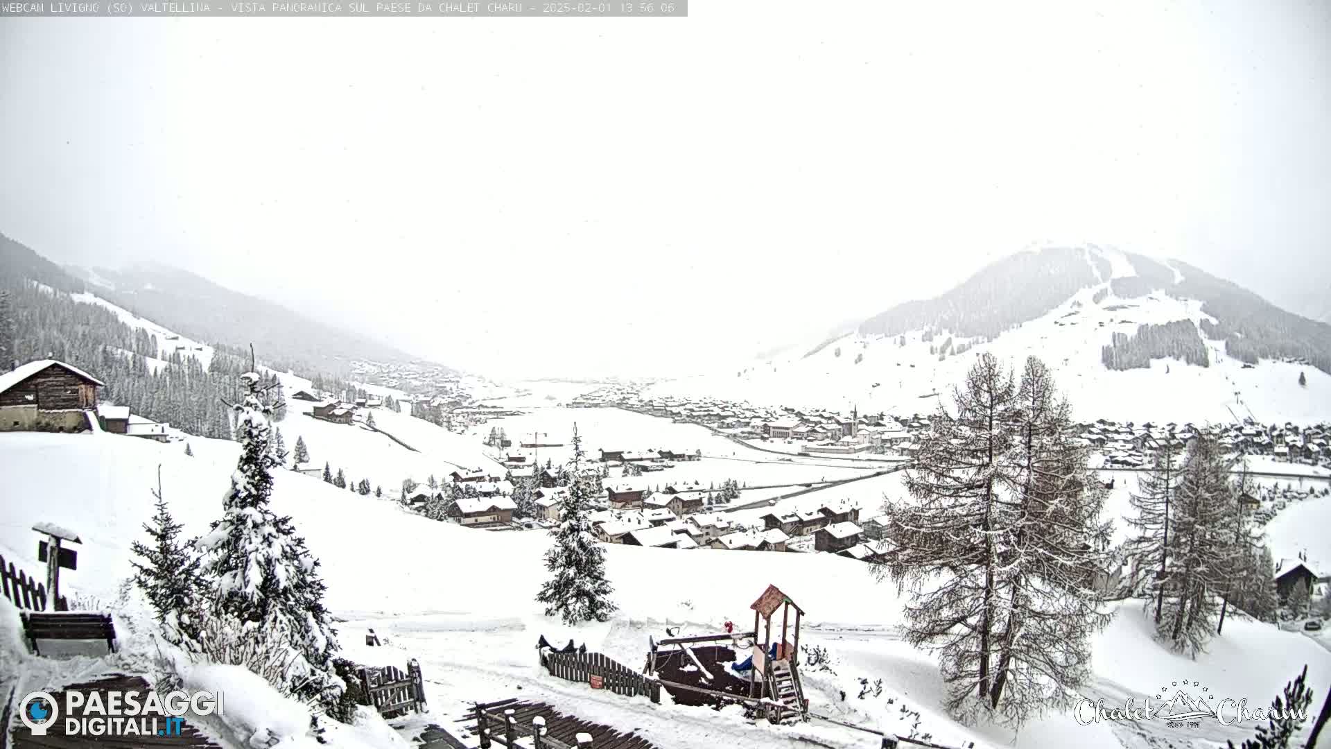 A snow-covered village nestled in a valley between snow-dusted mountains, under a heavy, overcast sky with light snowfall.