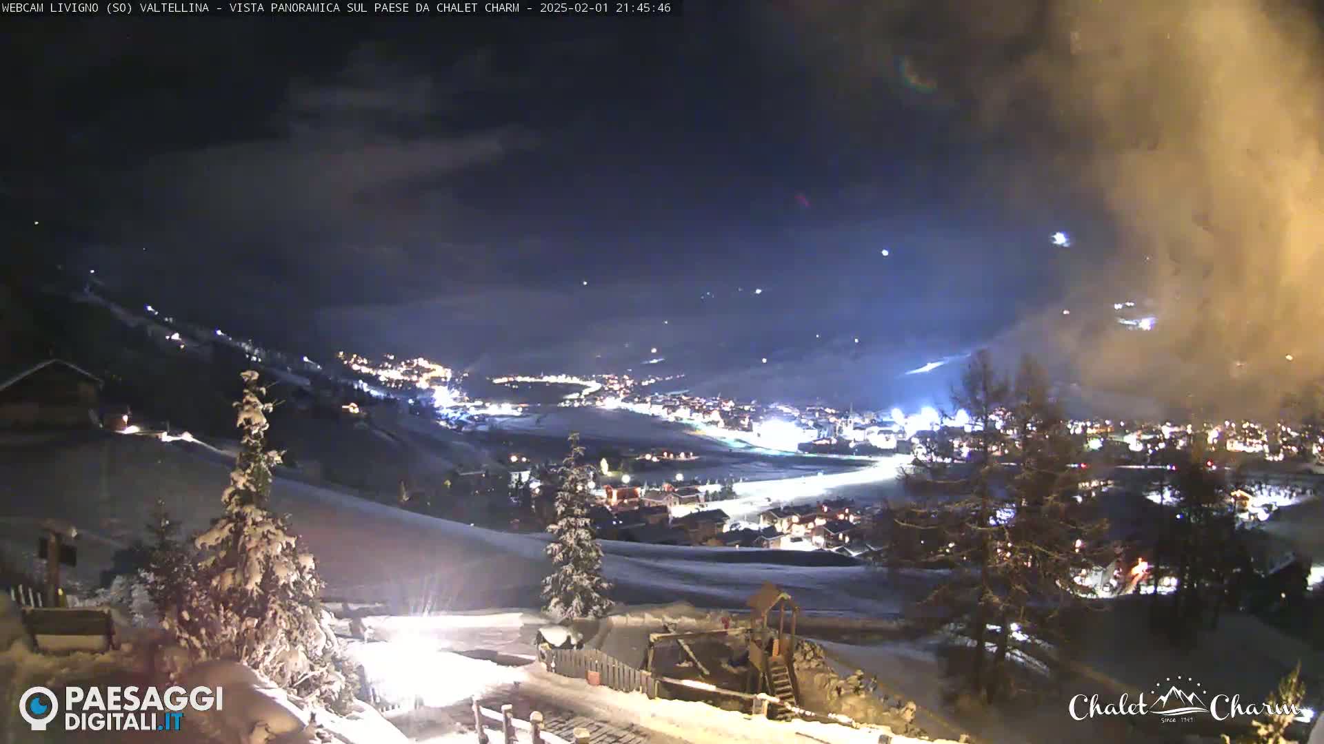 A snow-covered landscape at night, showing a town illuminated below, with a partly cloudy sky above.